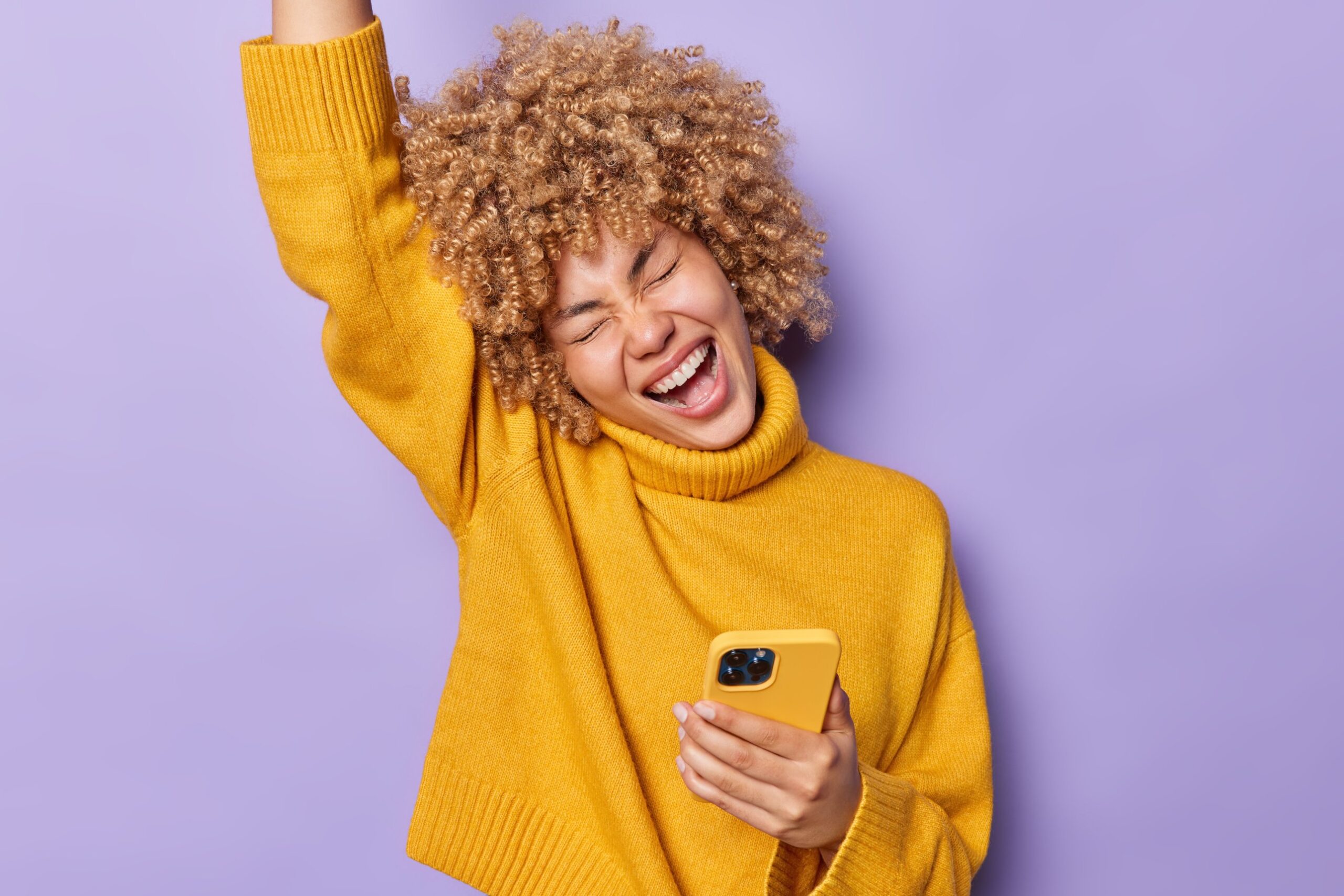 A woman in a yellow sweater laughing and raising her arm in celebration while holding her phone