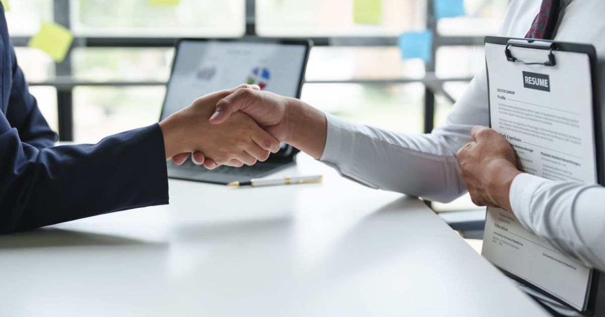Hiring manager shaking hands with a candidate holding a resume across an office table