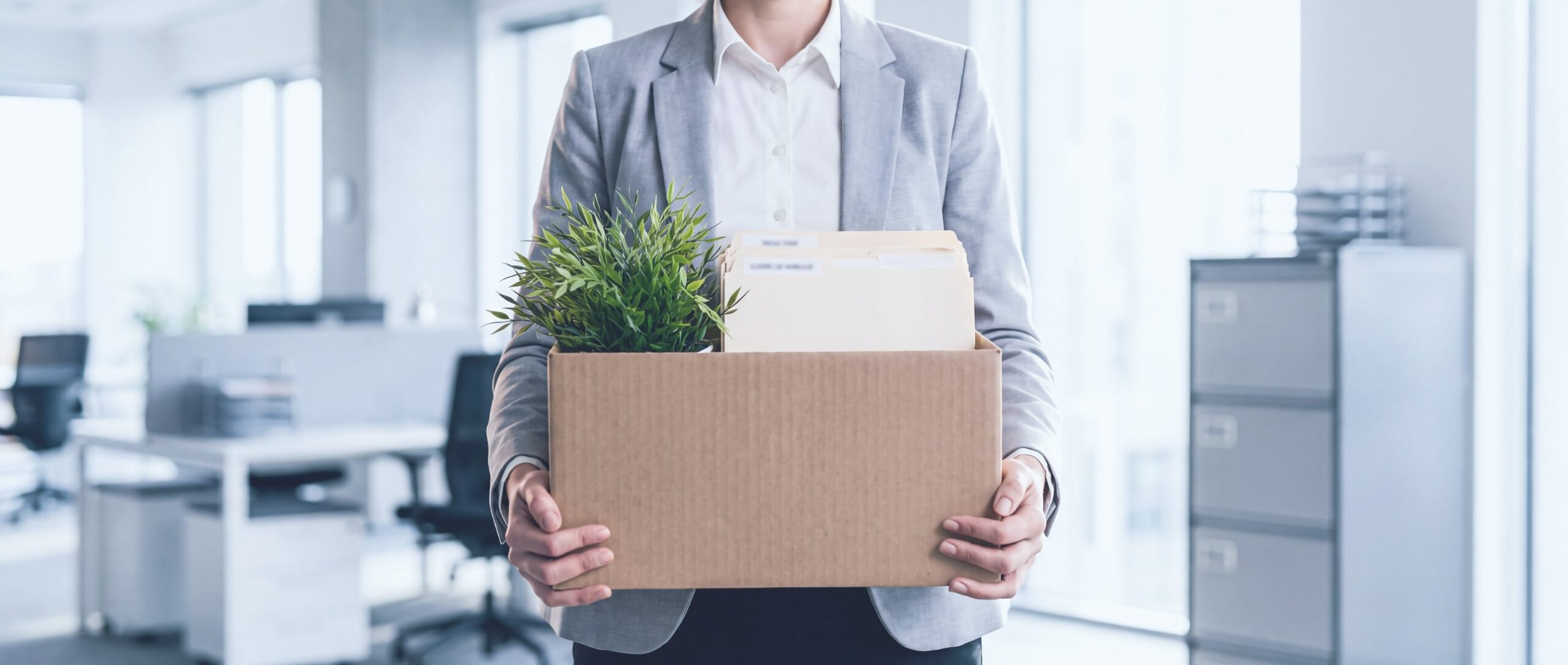 An employee in a gray blazer carrying a box with a plant and files out of the office