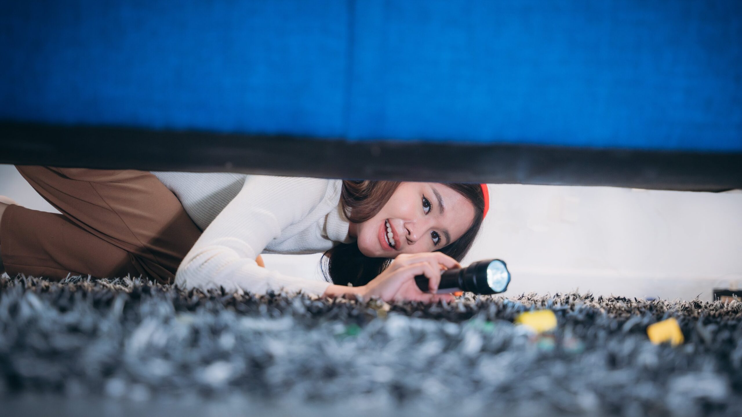 A woman lying on the floor shining a flashlight under a couch to see what she can find