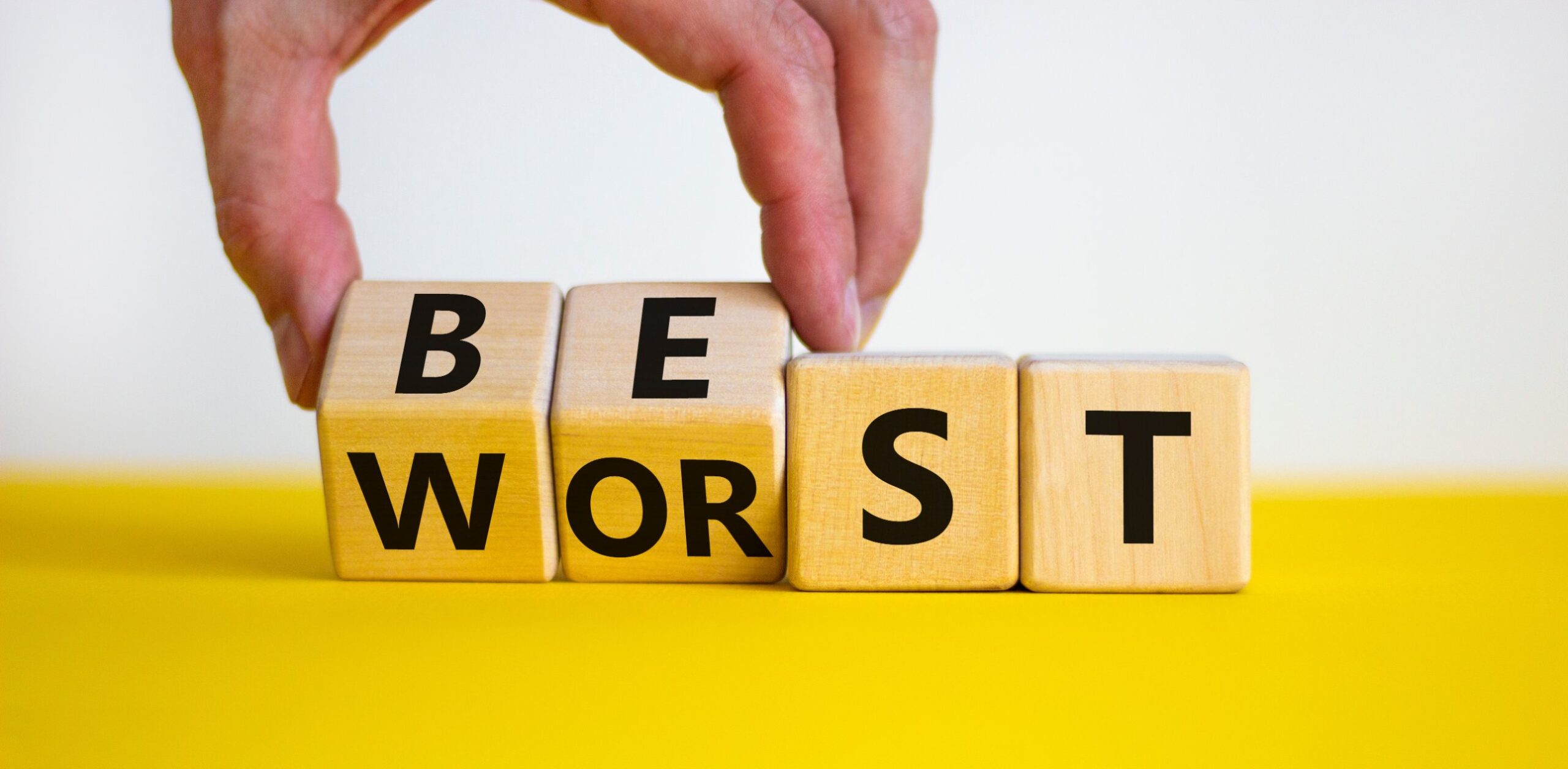 A hand flipping wooden blocks that spell WORST into BEST on a yellow surface