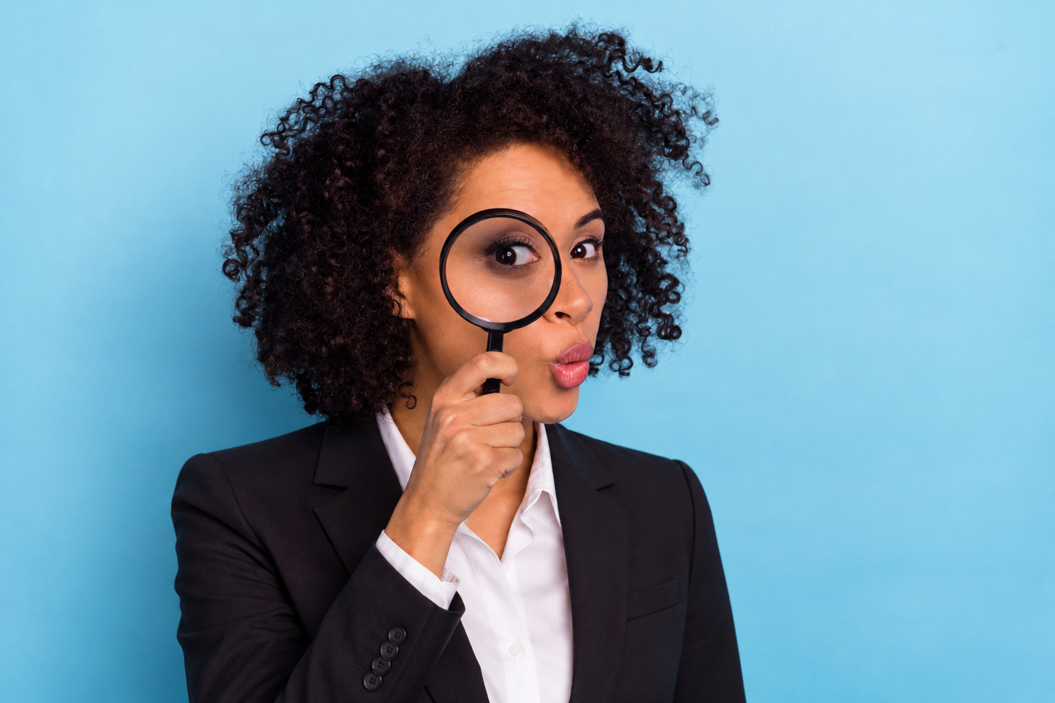 A professional woman in a black blazer looking closely through a magnifying glass